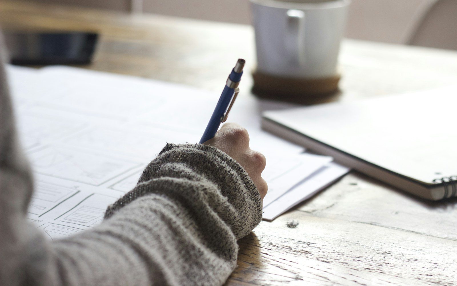 Person writing notes beside a laptop and coffee on a desk